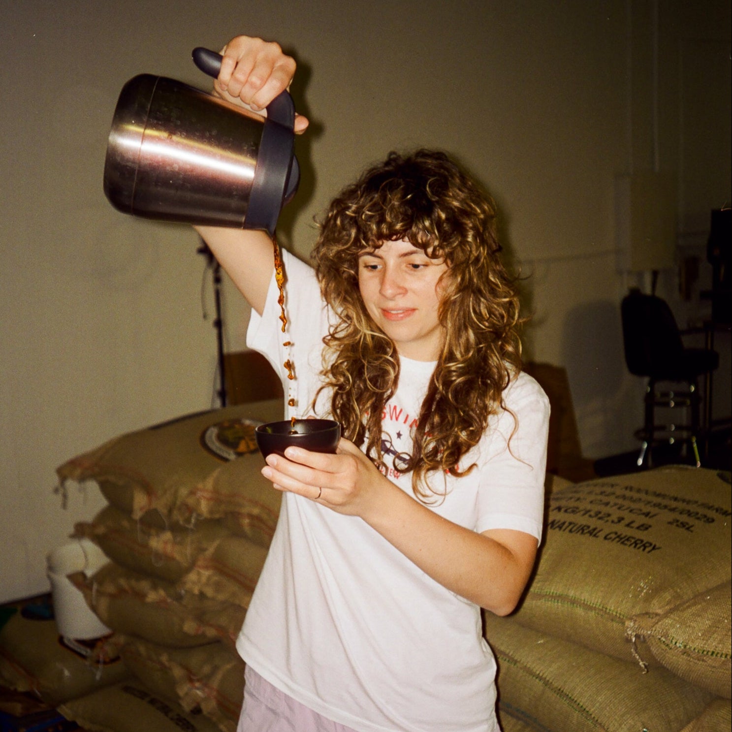 Film image of a person pouring coffee into a cup in an indoor setting with jute bags of coffee in the background.