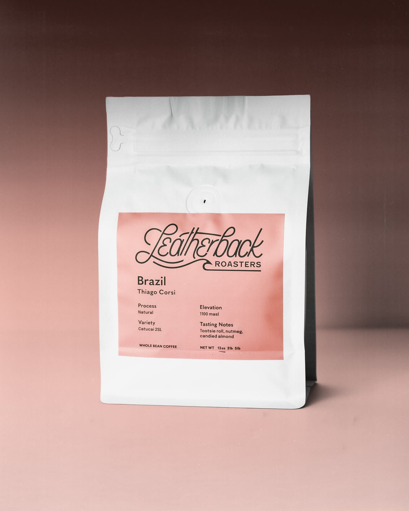A 12oz white valved coffee bag with a large pink label reading Leatherback Roasters and Brazil Thiago Corsi, photographed against a soft pink-to-brown gradient background.