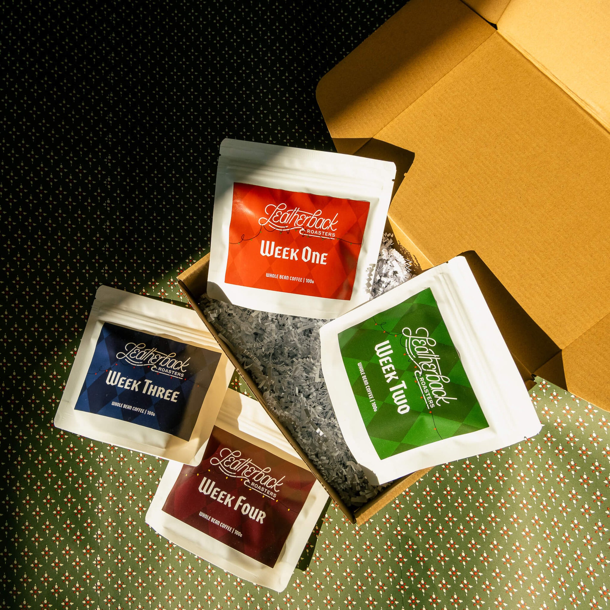 Advent Calendar Coffee Countdown 2025. 4 white bags of coffee with colorful Christmas-themed labels on a festive green patterned wrapping paper background.