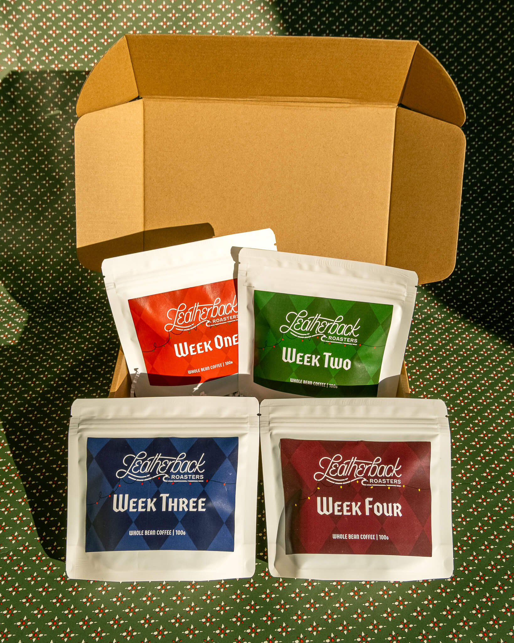 Advent Calendar Coffee Countdown 2025. 4 white bags of coffee with colorful Christmas-themed labels, in a kraft brown box on a festive green patterned wrapping paper background.