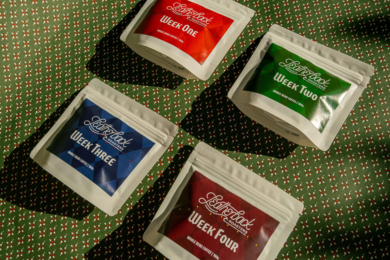 Advent Calendar Coffee Countdown 2025. 4 white bags of coffee with colorful Christmas-themed labels on a festive green patterned wrapping paper background.