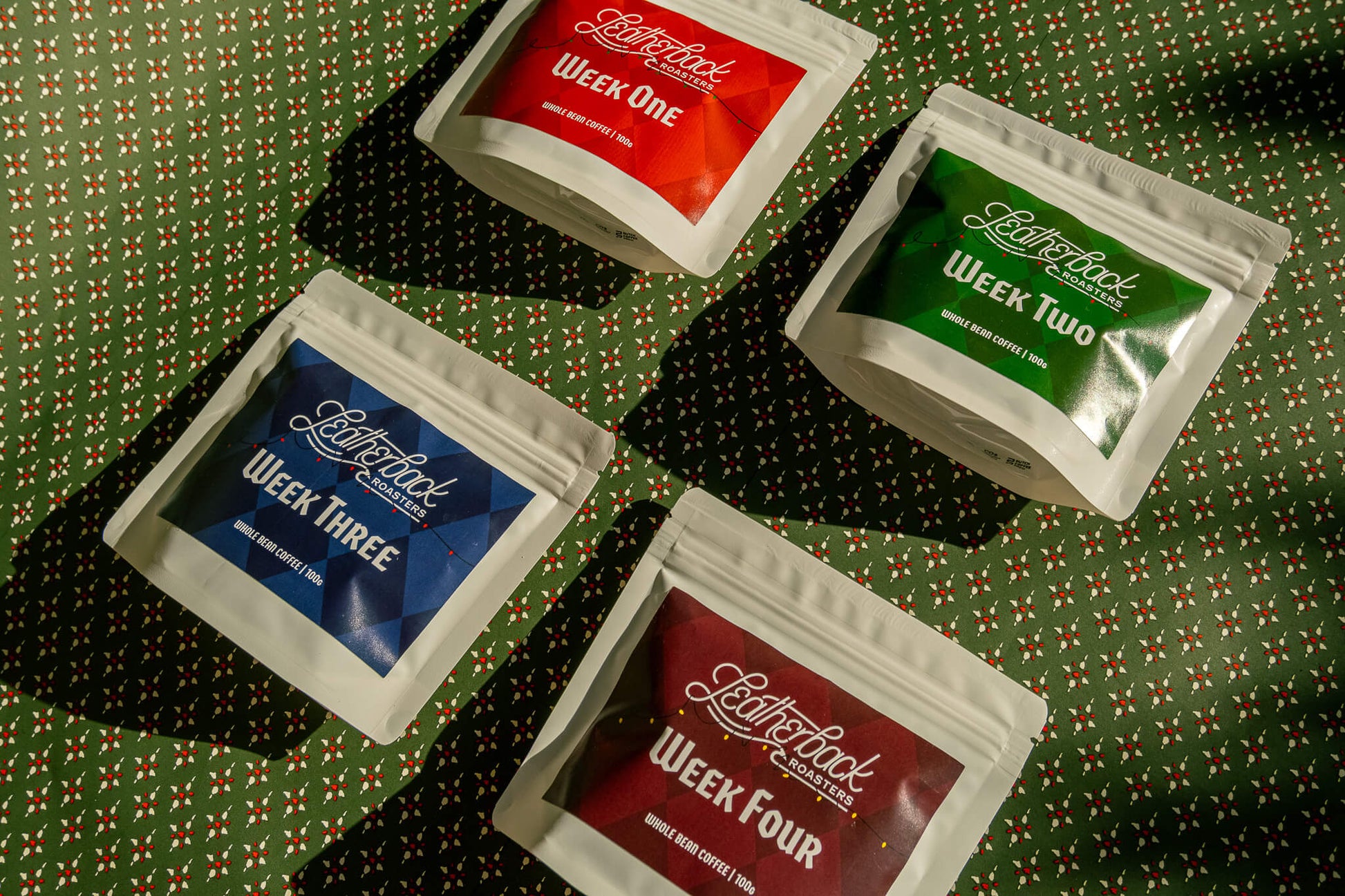 Advent Calendar Coffee Countdown 2025. 4 white bags of coffee with colorful Christmas-themed labels on a festive green patterned wrapping paper background.