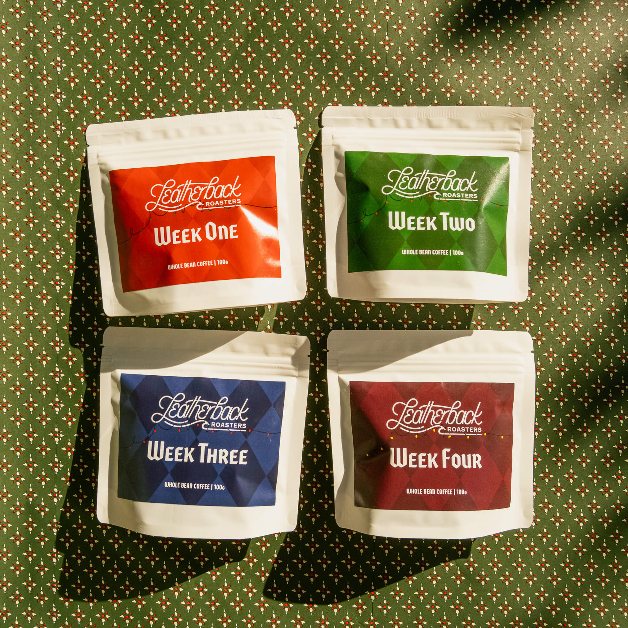 Advent Calendar Coffee Countdown 2025. 4 white bags of coffee with colorful Christmas-themed labels on a festive green patterned wrapping paper background.