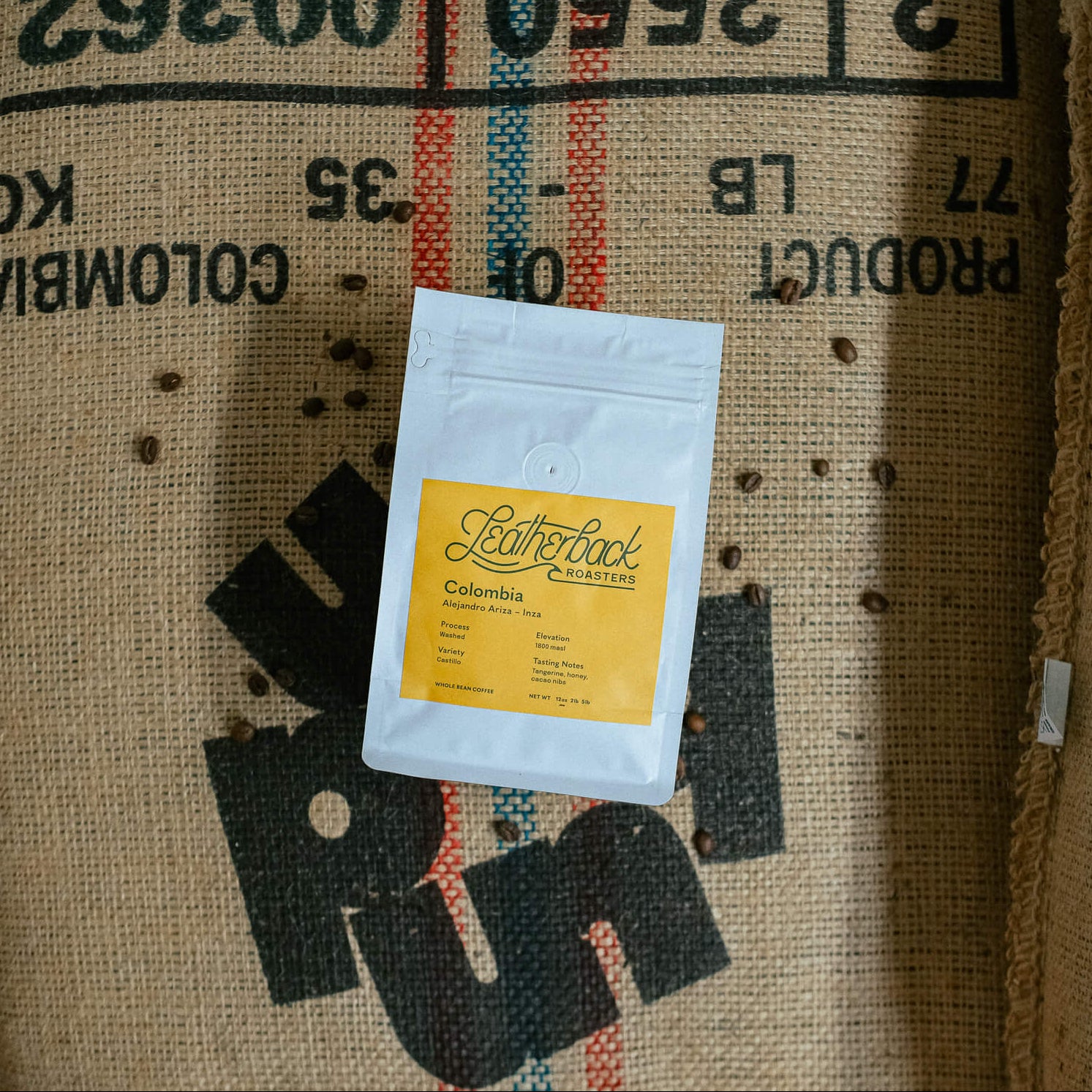 A white stand-up pouch with a yellow label reading 'Leatherback Coffee' and 'Colombia' lies on top of a large burlap sack printed with coffee origin text.