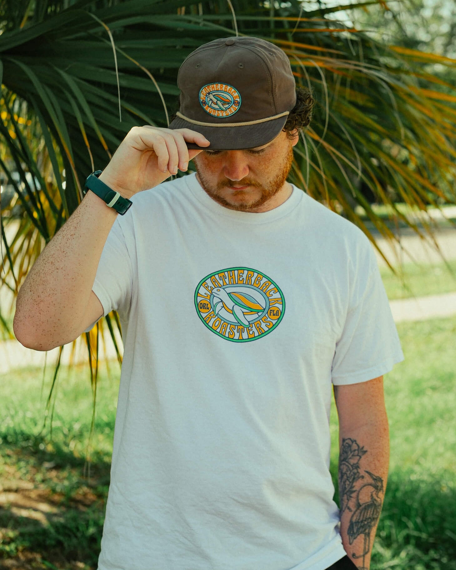 Man wearing a white t-shirt with a logo and a brown cap, standing outdoors with greenery in the background.