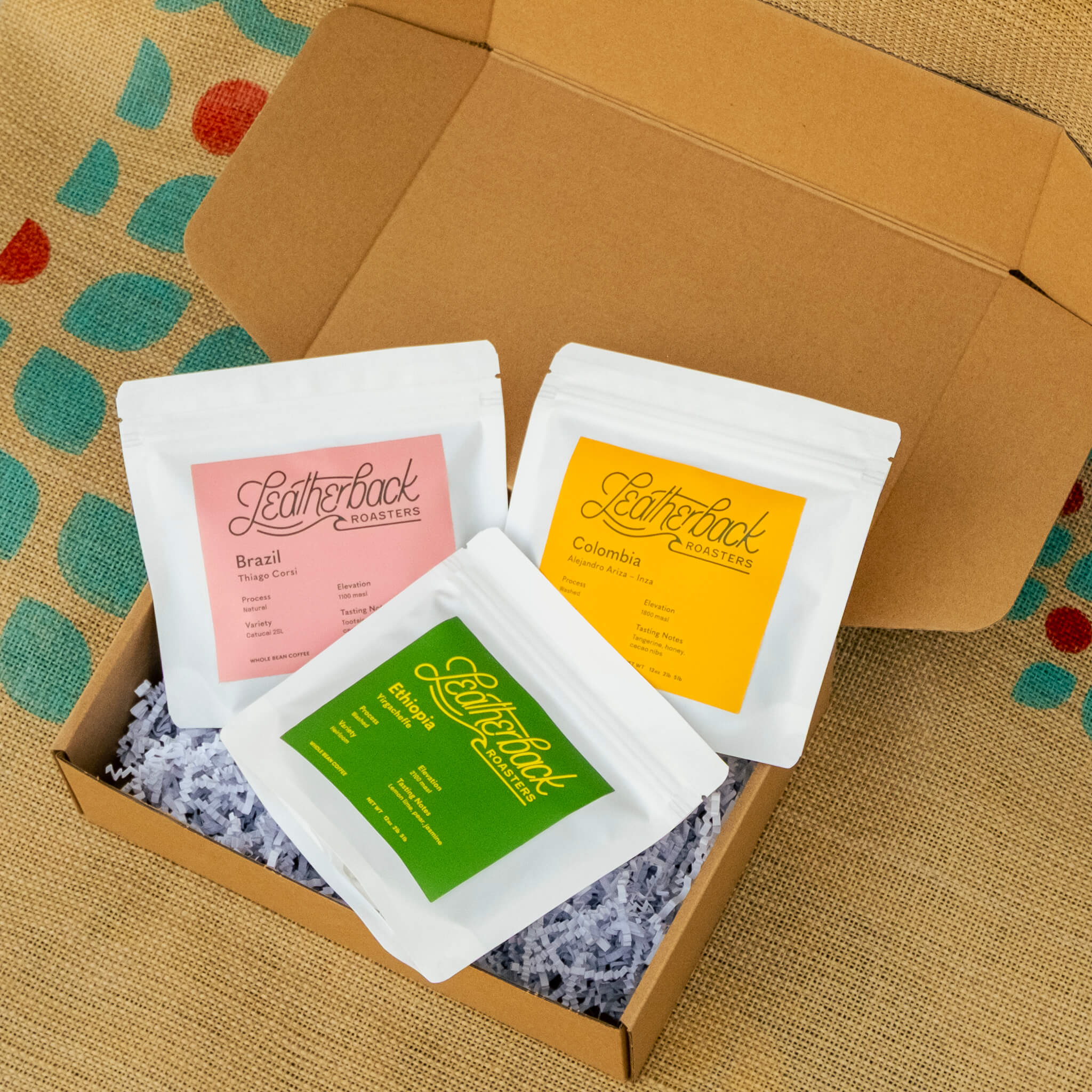 Three coffee packets with visible branding in a cardboard box on a jute bag.