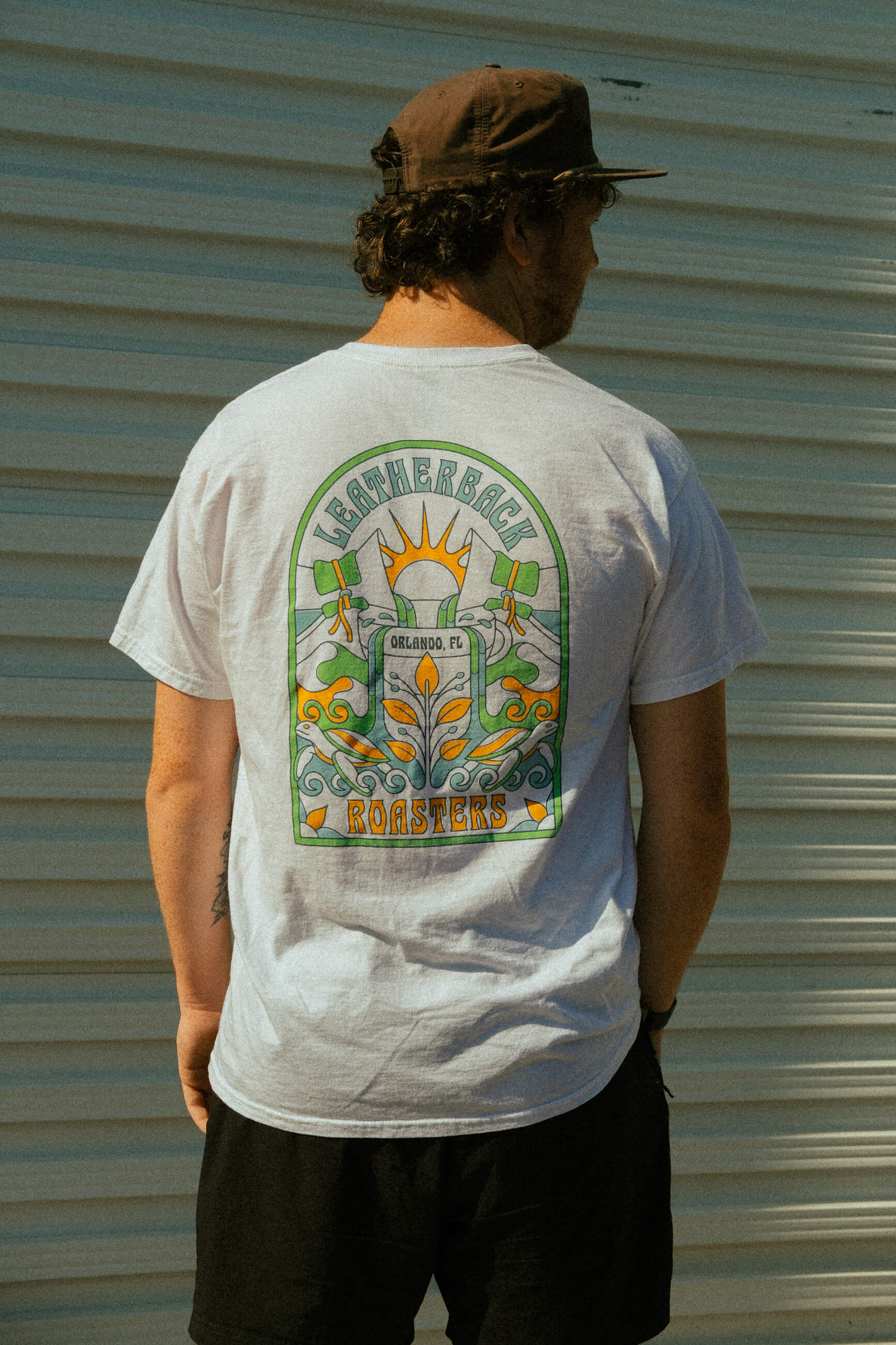 Person wearing a white t-shirt with a colorful graphic design on the back, standing against a corrugated metal wall.