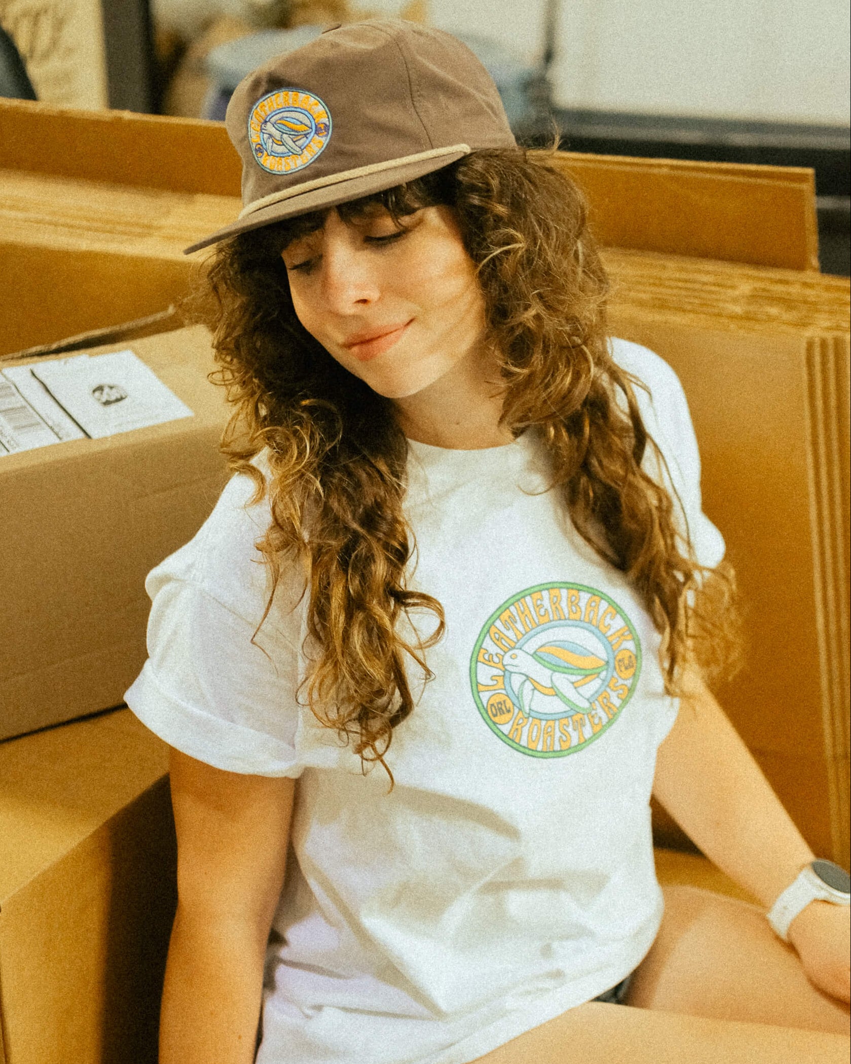Woman wearing a white t-shirt with a logo and a brown cap, sitting among cardboard boxes.
